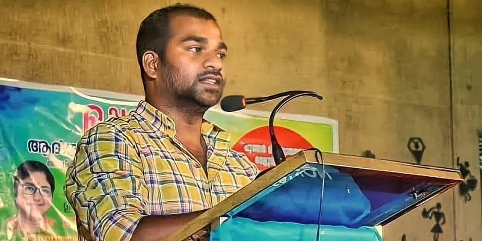 ചെക്ക് ഷർട്ട് ധരിച്ച മണികണ്ഠൻ സി പ്രസംഗപീഠത്തിൽ നിന്ന് മൈക്രോഫോണിൽ സംസാരിക്കുന്നു; അദ്ദേഹത്തിന് പിന്നിൽ മലയാളത്തിൽ അച്ചടിച്ച ഒരു ബാനറുണ്ട്.