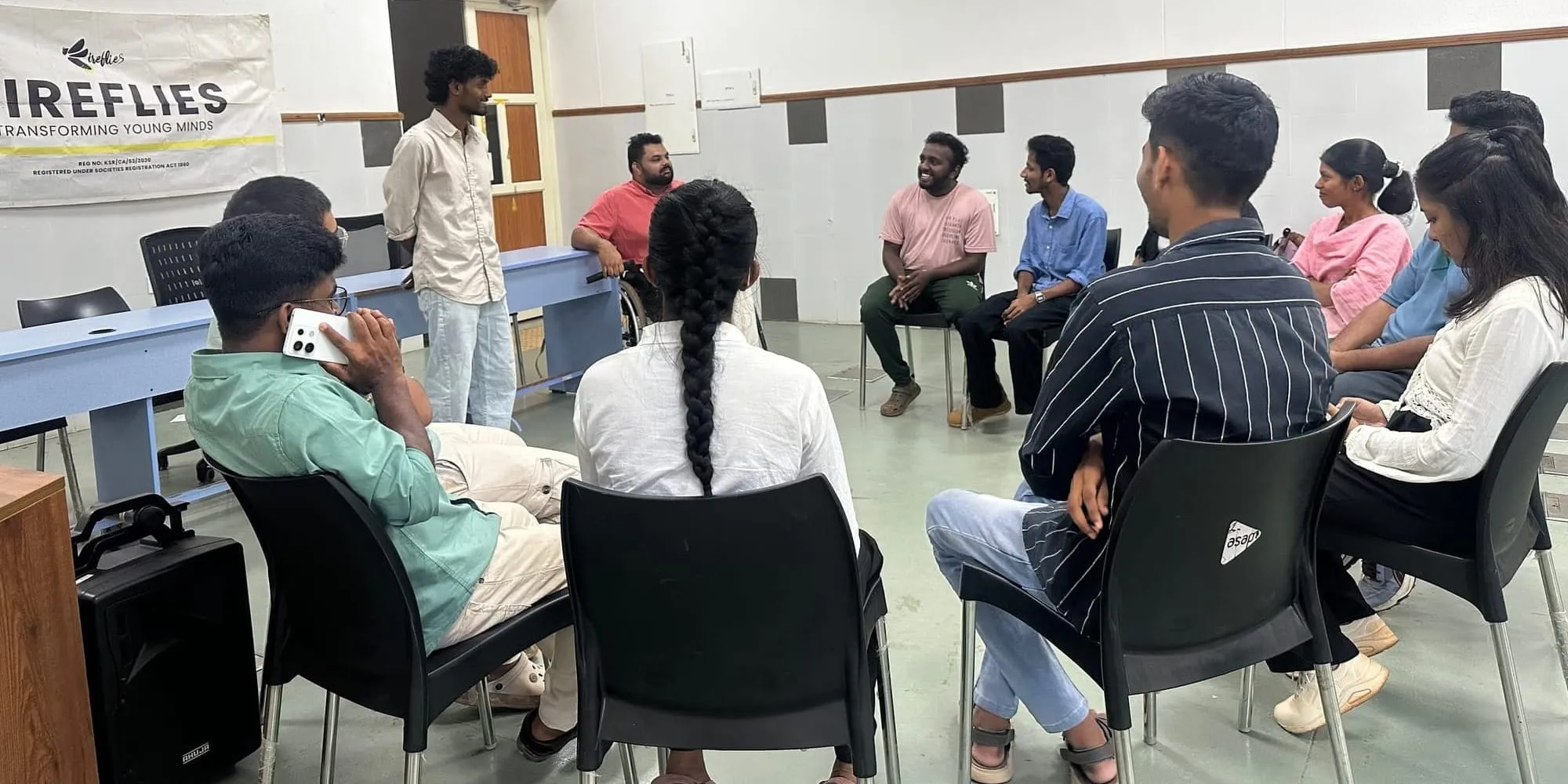 വർക്ക്ഷോപ്പ് മുറിയിൽ കറുത്ത കസേരകളുടെ വട്ടത്തിൽ ചെറുപ്പക്കാർ ഇരിക്കുമ്പോൾ ഒരാൾ എഴുന്നേറ്റുനിന്ന് സംസാരിക്കുന്നു; അദ്ദേഹത്തിന് പിന്നിലെ ബാനറിൽ "ഫയർഫ്ലൈസ് — ട്രാൻസ്ഫോർമിംഗ് യങ് മൈൻഡ്സ്" എന്ന് എഴുതിയിരിക്കുന്നു.