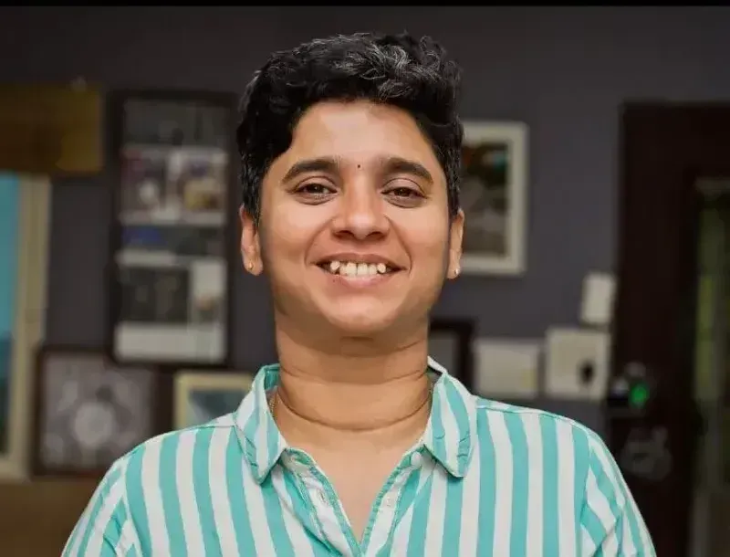 Ramya Sundararajan smiles at the camera in a teal-and-white striped shirt, framed pictures on the wall behind her.