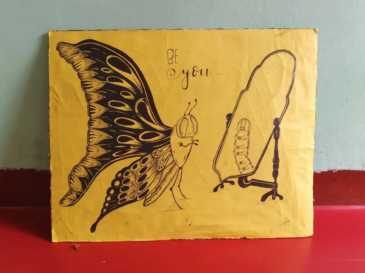 A large drawing on yellow paper propped against a wall shows a moth with patterned wings and a chrysalis on a stand, with the handwritten words "Be you".