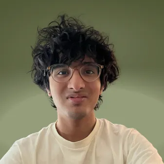 Arvind Unnithan, in round glasses and a cream T-shirt, photographed head and shoulders against a plain olive-green background.