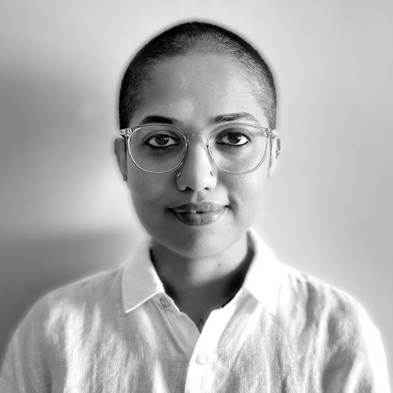Black-and-white portrait of Shradha in round glasses and a white collared shirt, close-cropped hair, against a plain grey background.
