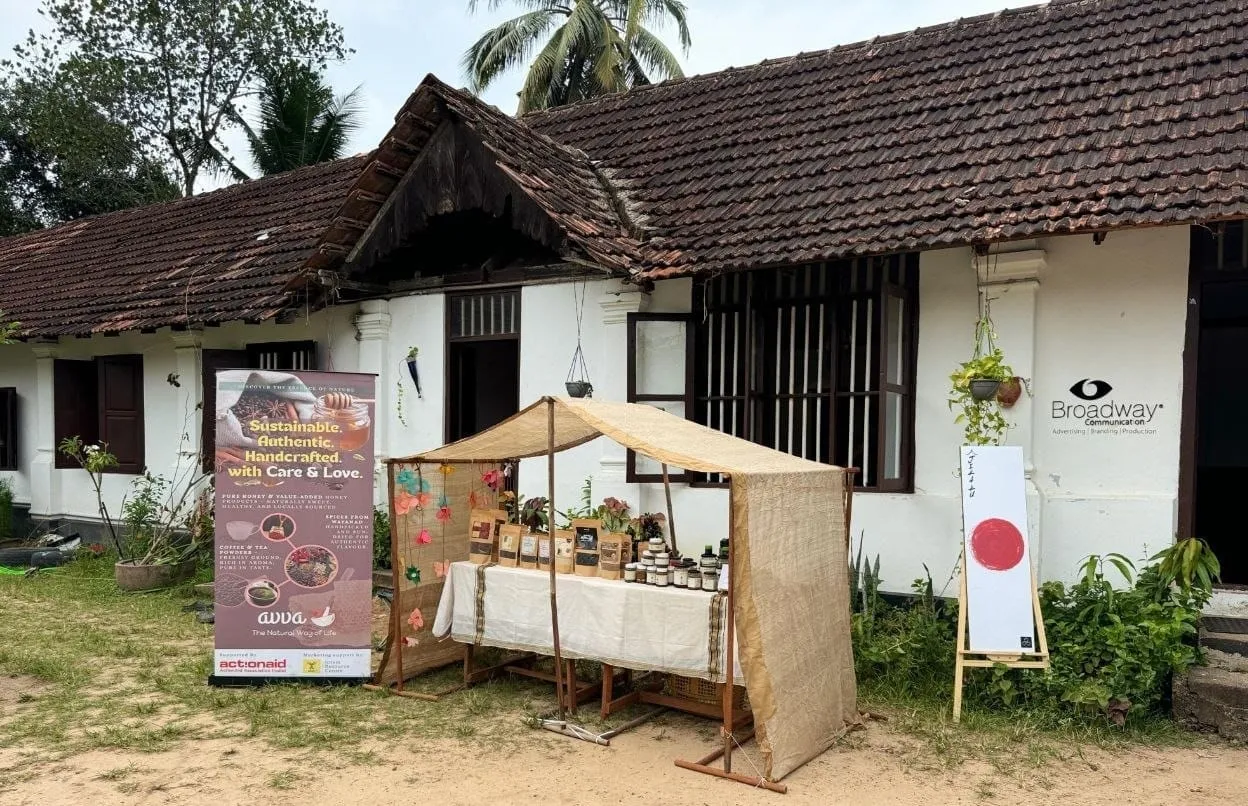 ഓടുമേഞ്ഞ വെളുത്ത വീടിനു മുന്നിലെ മണലിൽ സജ്ജീകരിച്ച, തുണി മേൽക്കൂരയുള്ള ഭരണികളുടെയും ഉൽപ്പന്നങ്ങളുടെയും ഒരു സ്റ്റാൾ; അതിനടുത്ത് "സുസ്ഥിരം. യഥാർത്ഥം. കൈകൊണ്ട് നിർമ്മിച്ചത്." എന്ന് എഴുതിയ ഒരു ബാനറുമുണ്ട്.