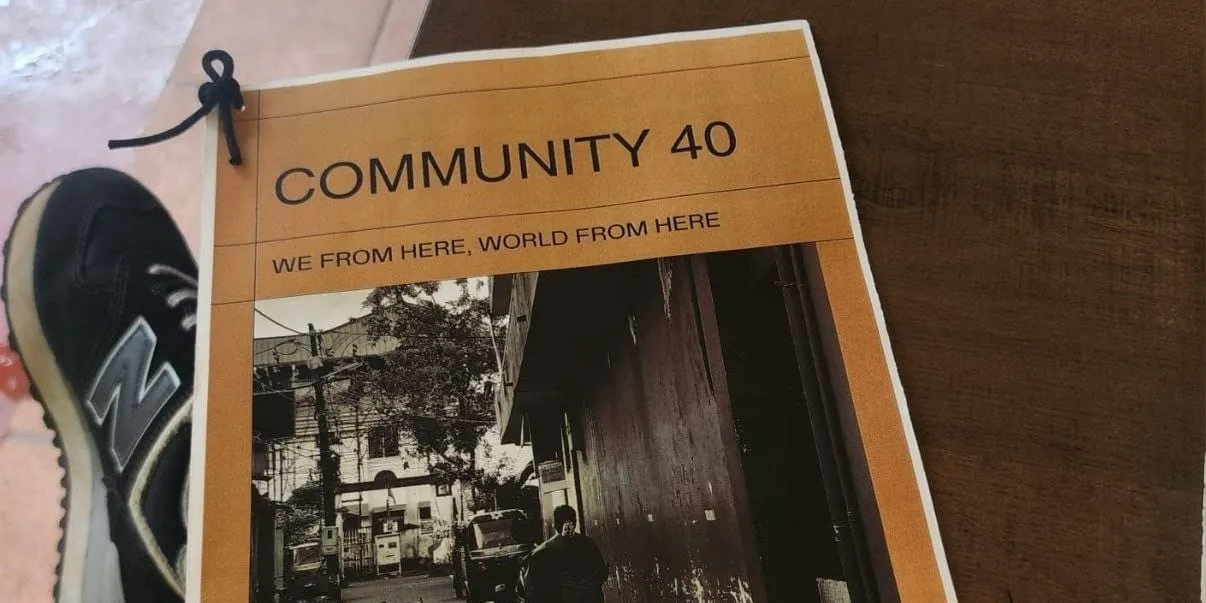 "കമ്മ്യൂണിറ്റി 40 — ഞങ്ങൾ ഇവിടെ നിന്ന്, ലോകം ഇവിടെ നിന്ന്" എന്ന തലക്കെട്ടിലുള്ള ഒരു ലഘുപുസ്തകം ട്രെയിനറുടെ അരികിലുള്ള കടും തവിട്ടുനിറത്തിലുള്ള മരമേശപ്പുറത്ത് ഇരിക്കുന്നു, അതിന്റെ കവറിൽ ഒരു കറുപ്പും വെളുപ്പും സ്ട്രീറ്റ് ഫോട്ടോഗ്രാഫുണ്ട്.