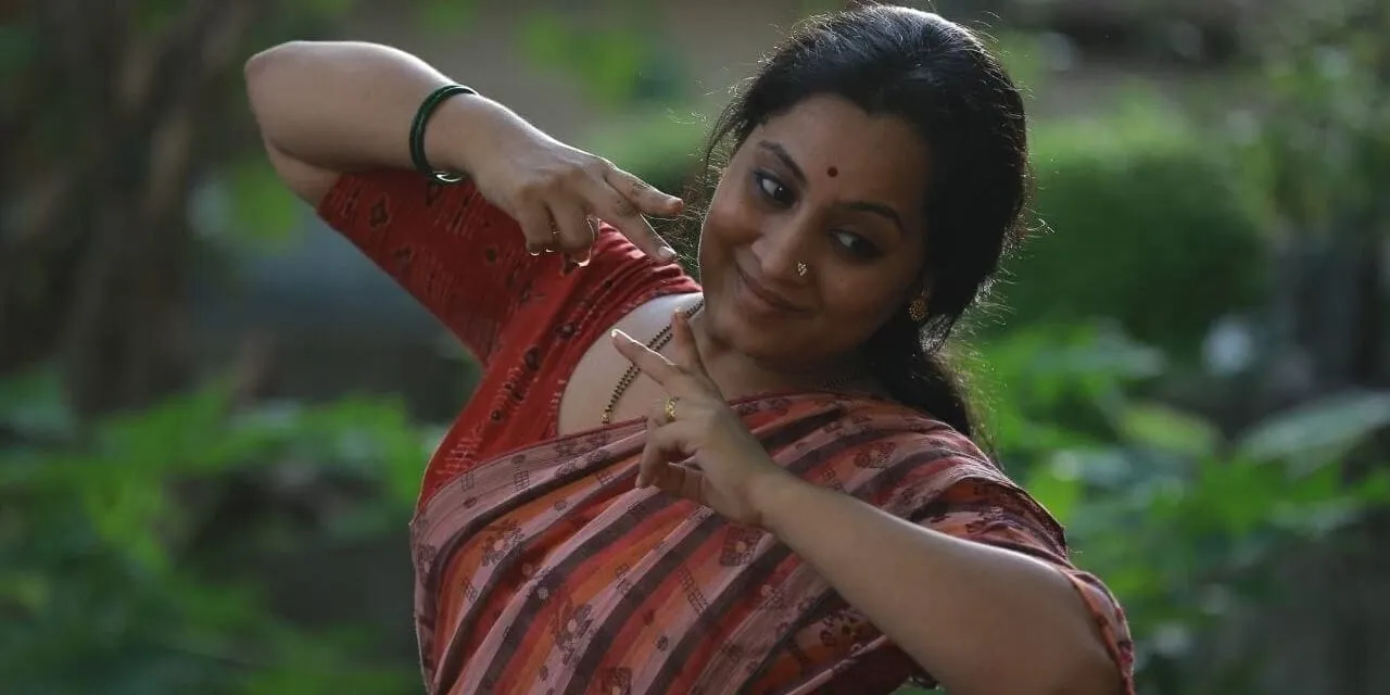 പൊന്നു സഞ്ജീവ് ചുവപ്പും ക്രീം നിറത്തിലുമുള്ള വസ്ത്രത്തിൽ, ഒരു കൈ തലയ്ക്കരികിൽ ഉയർത്തിയും മറ്റേ കൈ മുദ്രയിലും പിടിച്ച്, പശ്ചാത്തലത്തിൽ മങ്ങിയ പച്ചപ്പോടെ പുറത്ത് നൃത്തഭാവത്തിൽ നിൽക്കുന്നു.