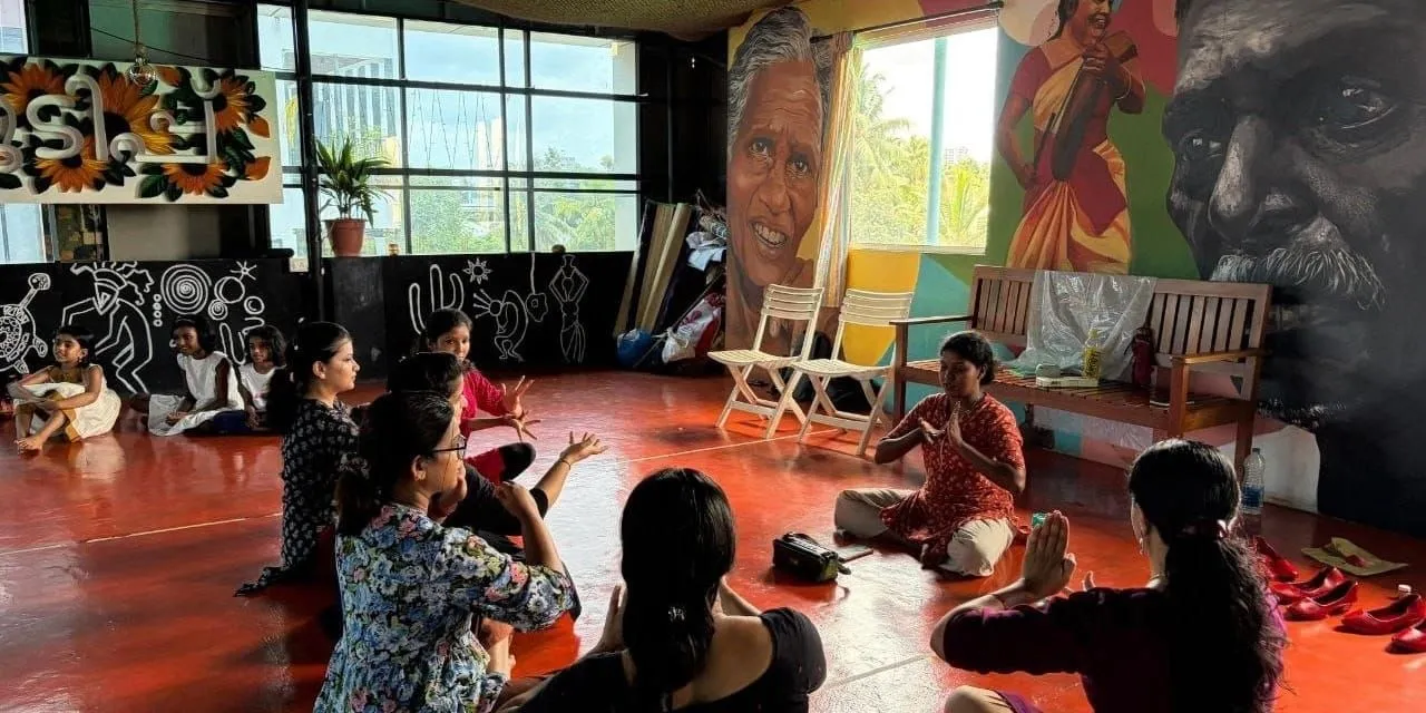 प्रतिभागी रेड ऑक्साइड के फर्श पर एक ढीले घेरे में बैठकर हाथ से इशारे कर रहे हैं, एक ऐसे हॉल में जहाँ चेहरों और एक संगीतकार के बड़े भित्तिचित्र बने हैं।