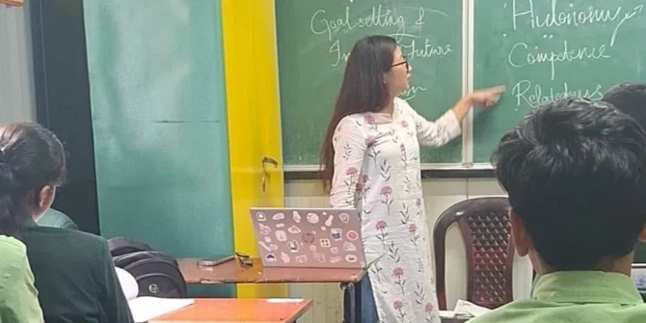 Sentinaro Shiu points to a chalkboard where "Goal setting", "Autonomy", "Competence" and "Relatedness" are written, students seated with their backs to the camera.