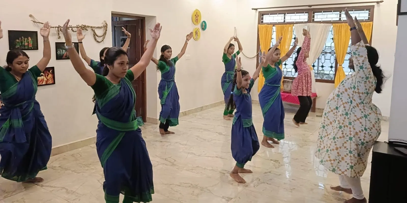 A row of dancers in blue and green practice saris hold the same raised-arm pose in a home studio, a teacher in a patterned kurta facing them.