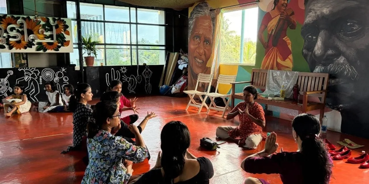 Participants sit in a loose circle on a red oxide floor making hand gestures, in a hall painted with large murals of faces and a musician.
