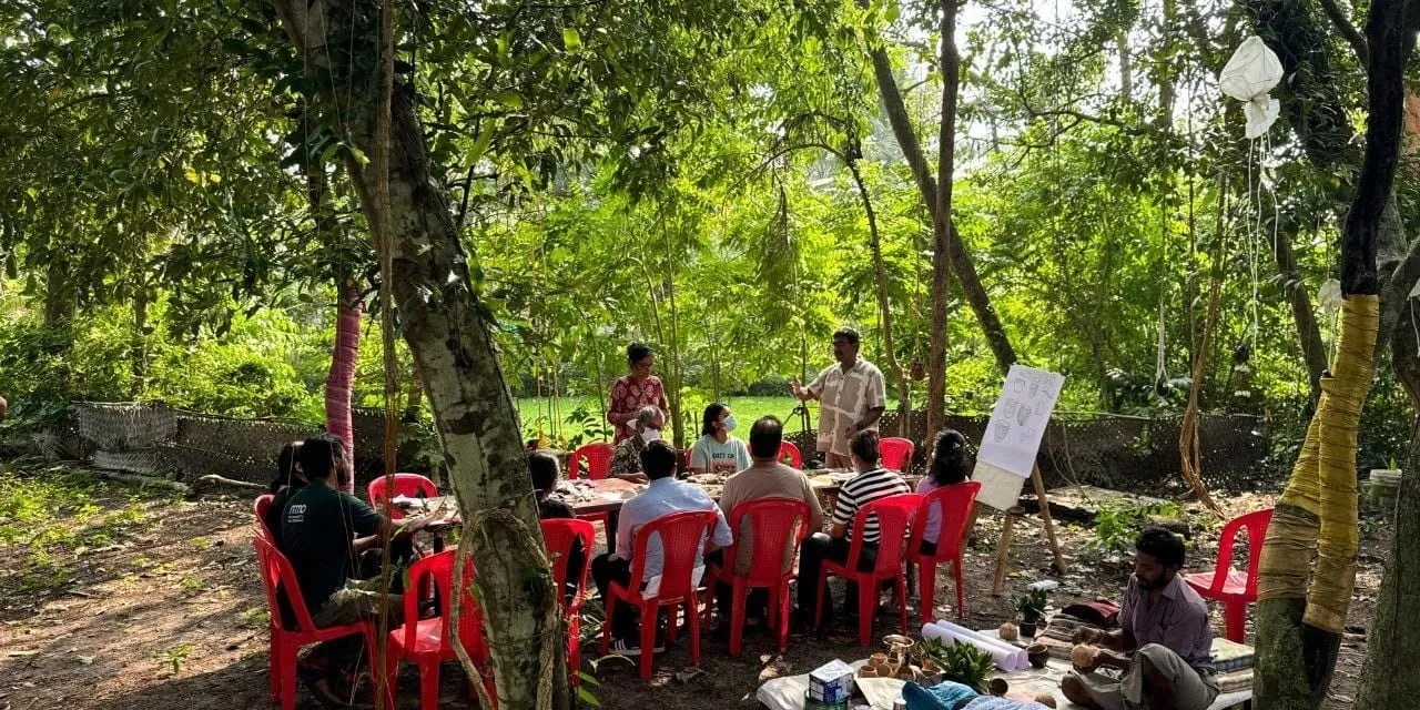 लोग पेड़ों के नीचे लाल प्लास्टिक की कुर्सियों की पंक्तियों में बैठे हैं, जो एक ईज़ल पर लगे फ़्लिप चार्ट के पास खड़े वक्ता का सामना कर रहे हैं, और पीछे एक हरा मैदान है।