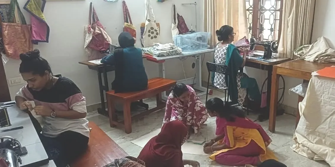 A tailoring workshop — women work at sewing machines along the far wall while others sit on the floor with fabric; finished cloth bags hang above them.