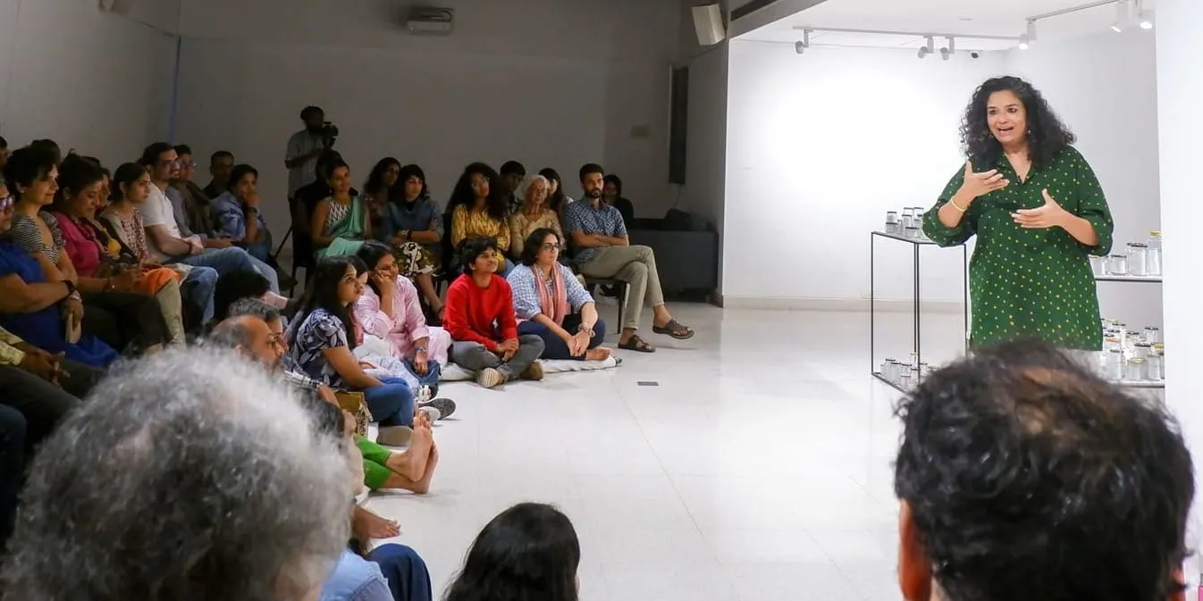 निमि रवींद्रन हरे रंग के पोल्का-डॉट कुर्ते में हाथ उठाए बोलती हुई खड़ी हैं, एक सफेद कमरे में कुर्सियों और फर्श पर दर्शक बैठे हैं।
