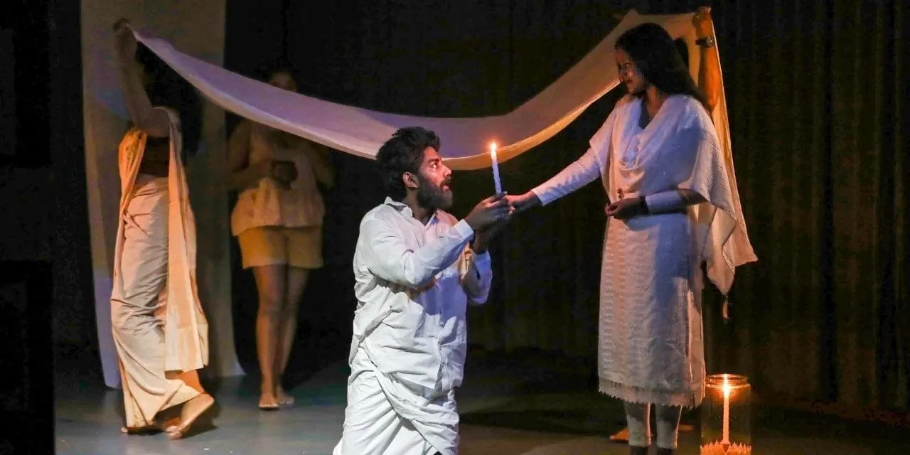 A stage scene in low light — a performer kneels holding a lit candle while another stands with an arm outstretched, a pale cloth stretched overhead.