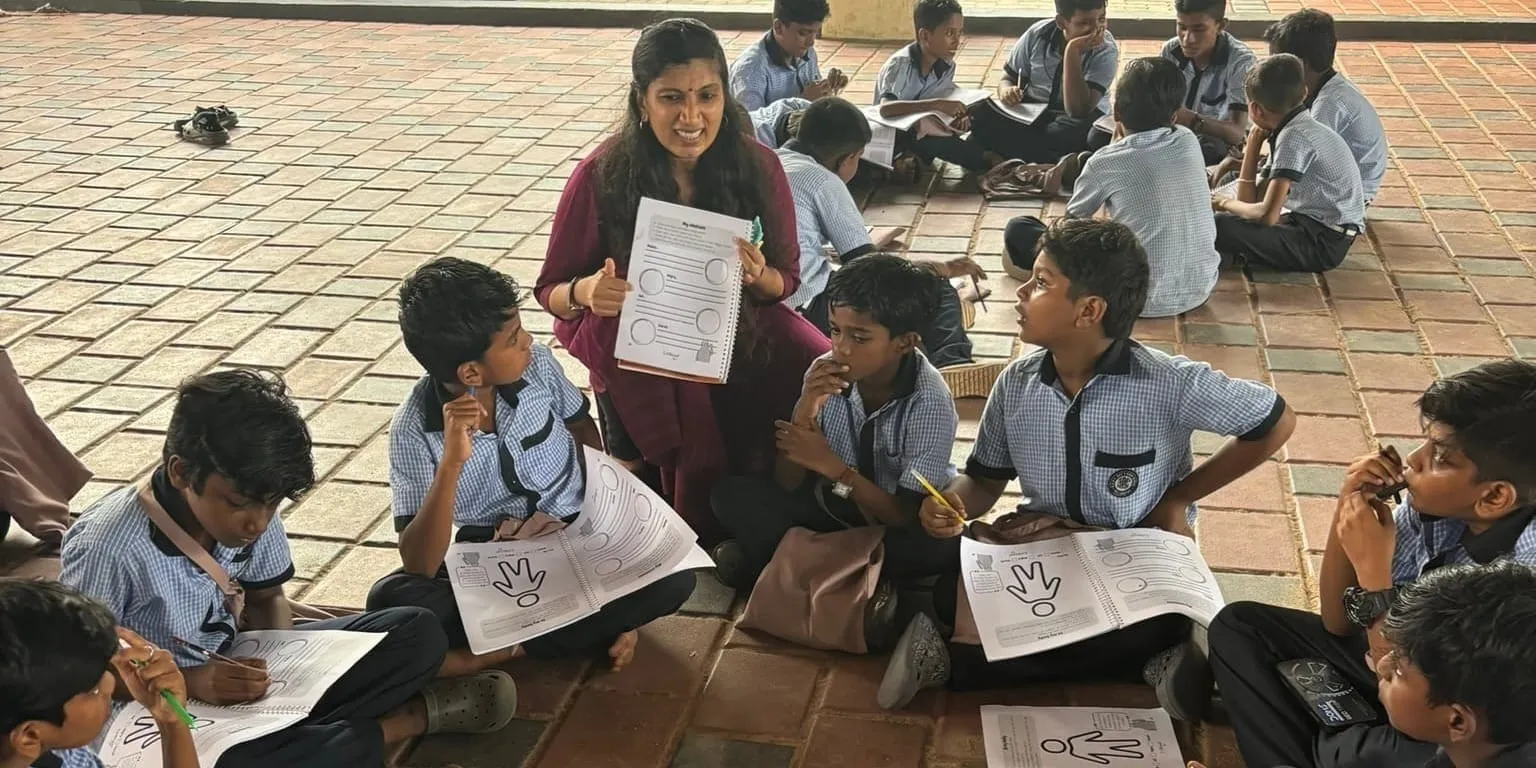 आरती वीरुपिल्लई चेक वाली यूनिफॉर्म पहने एक दर्जन स्कूली बच्चों के बीच टाइल वाले फर्श पर पालथी मारकर बैठी हैं और एक वर्कशीट हाथ में उठाए हुए हैं जबकि बच्चे अपनी-अपनी कॉपियां भर रहे हैं।