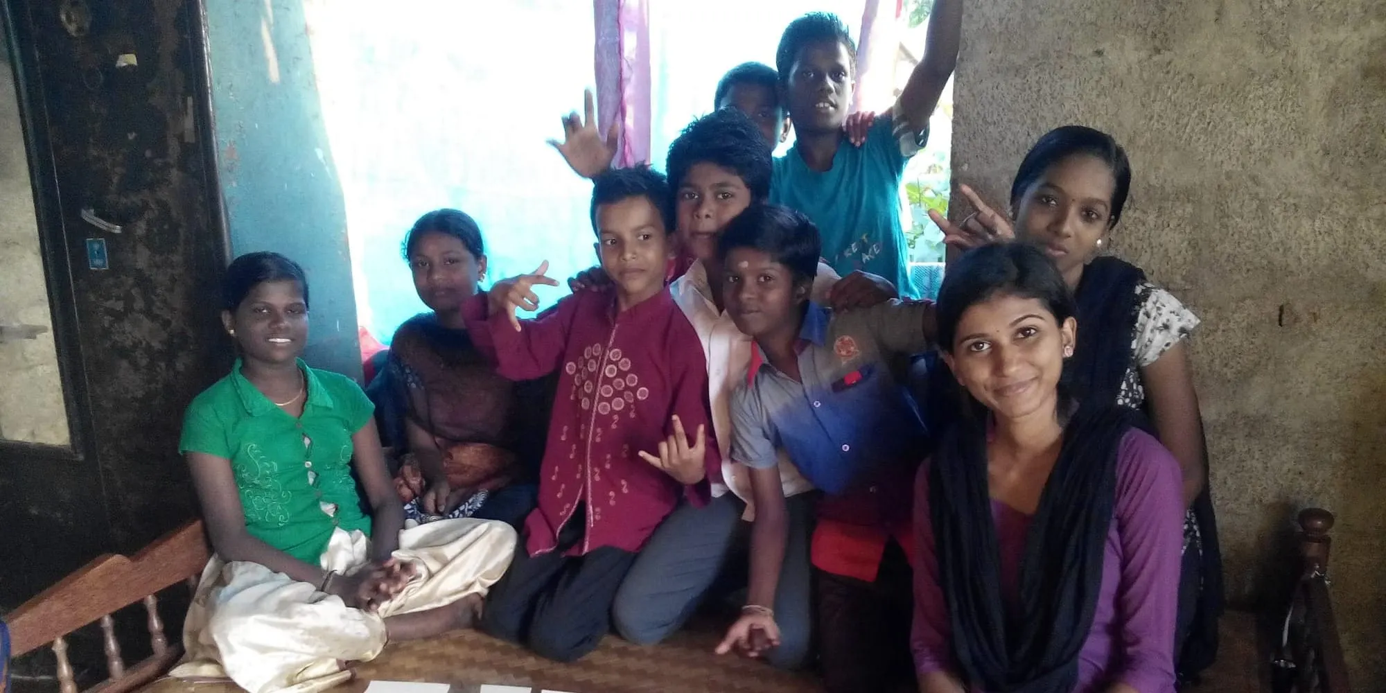 ഒരു ജാലകത്തിനരികിലെ മുറിയുടെ നിലത്ത് കുട്ടികളും യുവതികളും അടുത്തടുത്തിരിക്കുന്നു, ചിലർ ക്യാമറയിലേക്ക് കൈവീശുകയും സമാധാനചിഹ്നം കാണിക്കുകയും ചെയ്യുന്നു.