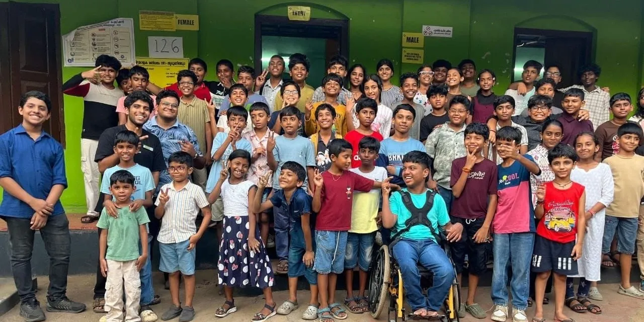 പച്ച ചുവരുകളുള്ള കെട്ടിടത്തിന് പുറത്ത് ഗ്രൂപ്പ് ഫോട്ടോയ്ക്കായി അമ്പതോളം കുട്ടികളും യുവ സന്നദ്ധപ്രവർത്തകരും ഒത്തുകൂടുന്നു, മുൻനിരയിലുള്ള ഒരു കുട്ടി വീൽചെയർ ഉപയോഗിക്കുന്നു.