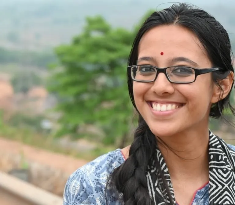 ശിലഞ്ജനി ഭട്ടാചാര്യ കണ്ണടയും നീലയും വെള്ളയും പാറ്റേണുള്ള ടോപ്പും ധരിച്ച് പുറത്ത് പുഞ്ചിരിച്ചുനിൽക്കുന്നു, മുടി പിന്നിയിട്ടിരിക്കുന്നു, പശ്ചാത്തലത്തിൽ മലനിരകൾ മങ്ങിയിരിക്കുന്നു.