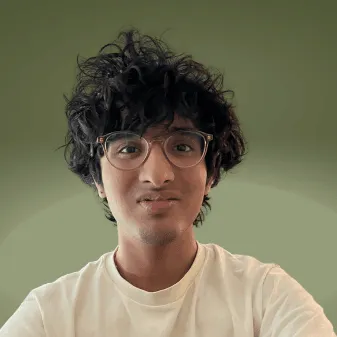 गोल चश्मे और क्रीम टी-शर्ट पहने अरविंद उन्निथन, एक सादे जैतून-हरे बैकग्राउंड के सामने ली गई सिर और कंधों तक की तस्वीर।