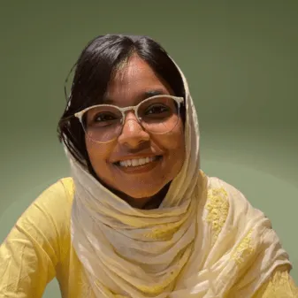 Haneen Naseer in a pale yellow headscarf and kurta, wearing glasses and smiling, photographed against an olive-green background.