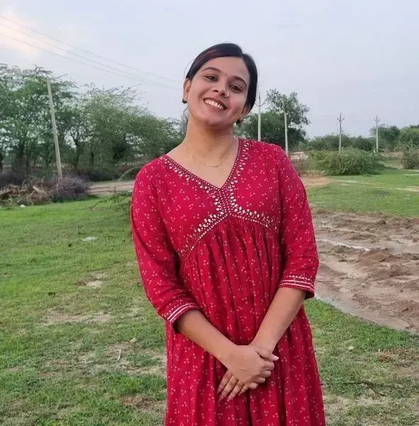 हृद्या झाड़ीदार घास पर लाल रंग के कढ़ाईदार कुर्ते में खड़ी है, उसके हाथ आगे की ओर बंधे हैं, और पृष्ठभूमि में पेड़ और बिजली की लाइनें दिखाई दे रही हैं।