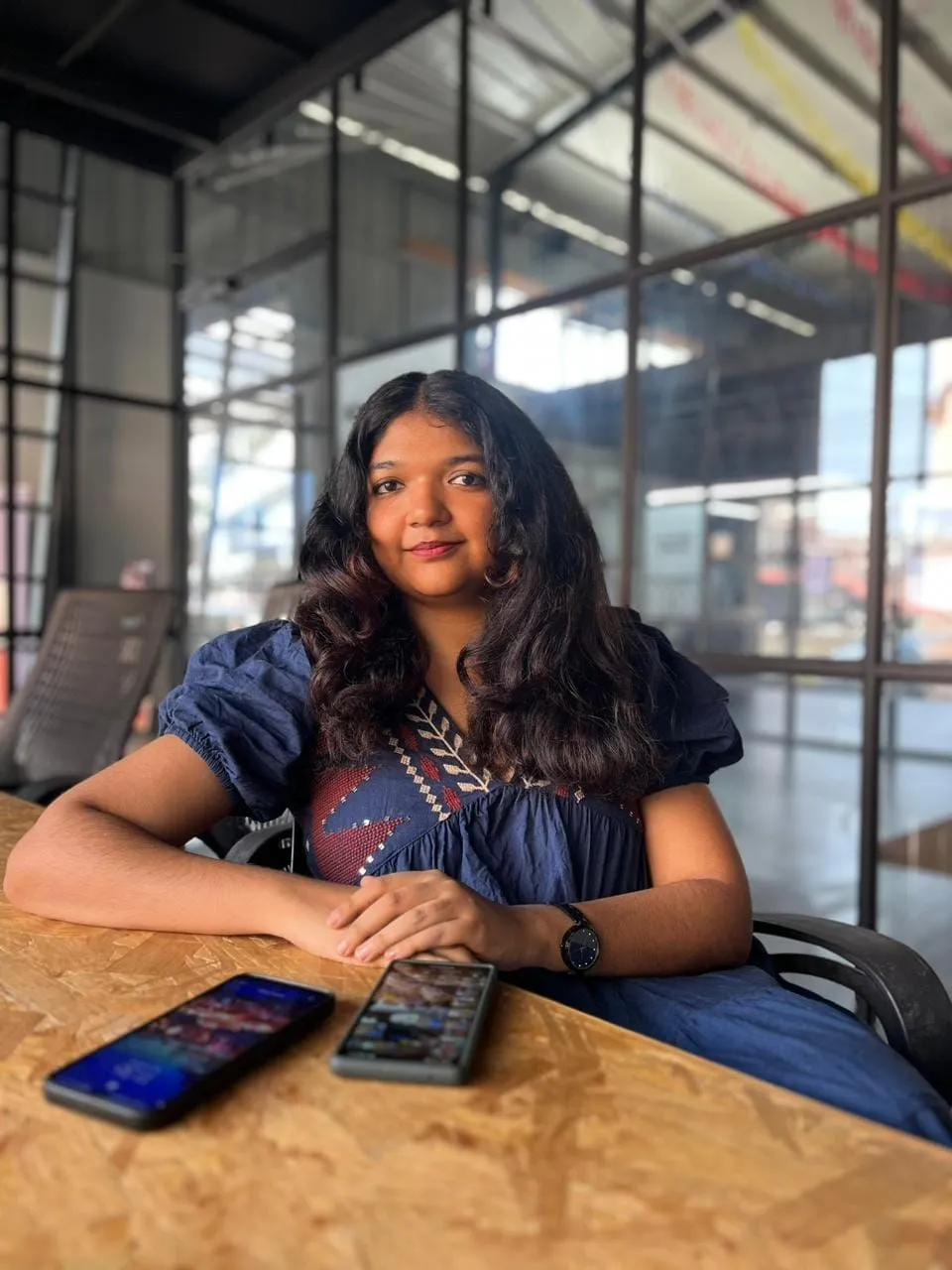 Arundhathi sits at a wooden table by a steel-framed window, arms folded in front of her, a phone and a small device on the table.