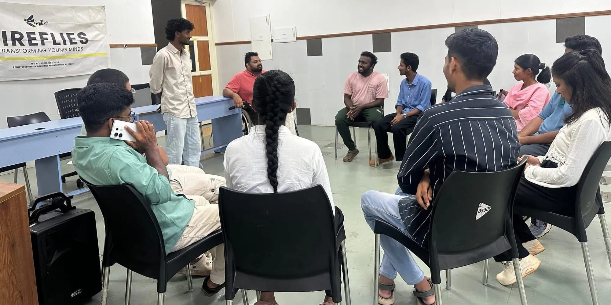 युवा एक कार्यशाला कक्ष में काली कुर्सियों के घेरे में बैठे हैं जबकि एक व्यक्ति खड़ा होकर बोल रहा है; उसके पीछे एक बैनर पर लिखा है "फ़ायरफ़्लाइज़ — ट्रांसफ़ॉर्मिंग यंग माइंड्स"।