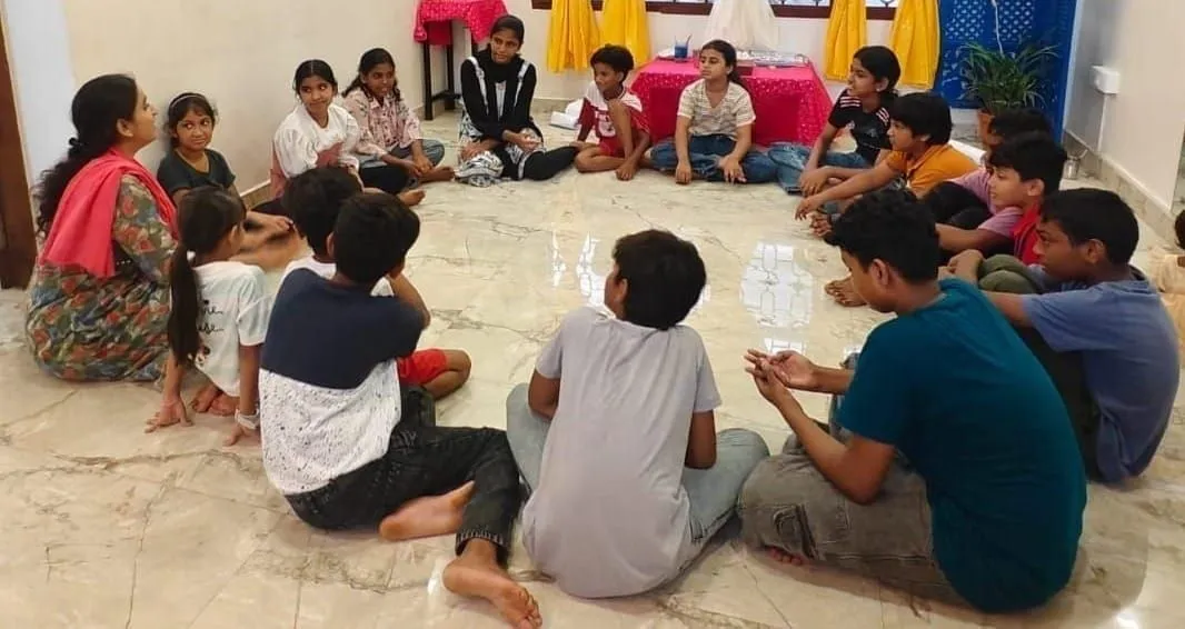 बच्चे संगमरमर के फर्श पर एक घेरे में बैठे हैं जिनके बीच कागज़ के छोटे कप हैं, घेरे के किनारे साड़ी में एक वयस्क महिला है।