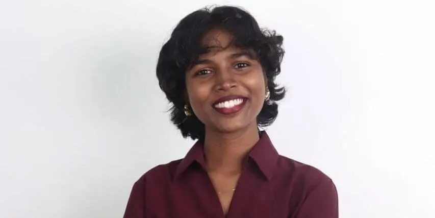 शिल्पा सुगुणन मैरून शर्ट में खुलकर मुस्कुरा रही हैं, जिनकी एक सादे हल्के रंग के बैकग्राउंड में सिर और कंधों की तस्वीर ली गई है।