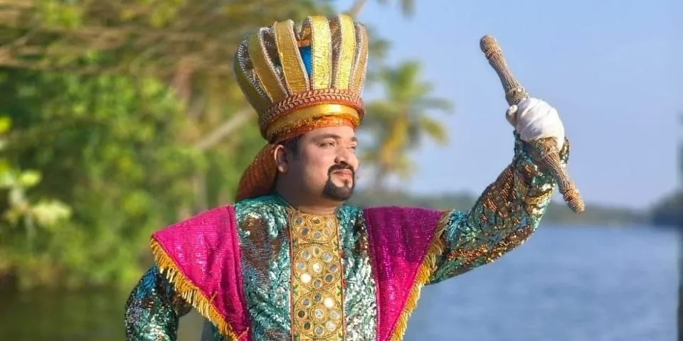 Anson Kurumbathuruth in full Chavittu Nadakam costume — a tall gold crown and a pink and turquoise sequinned cape — his sword raised, backwaters and palms behind him.