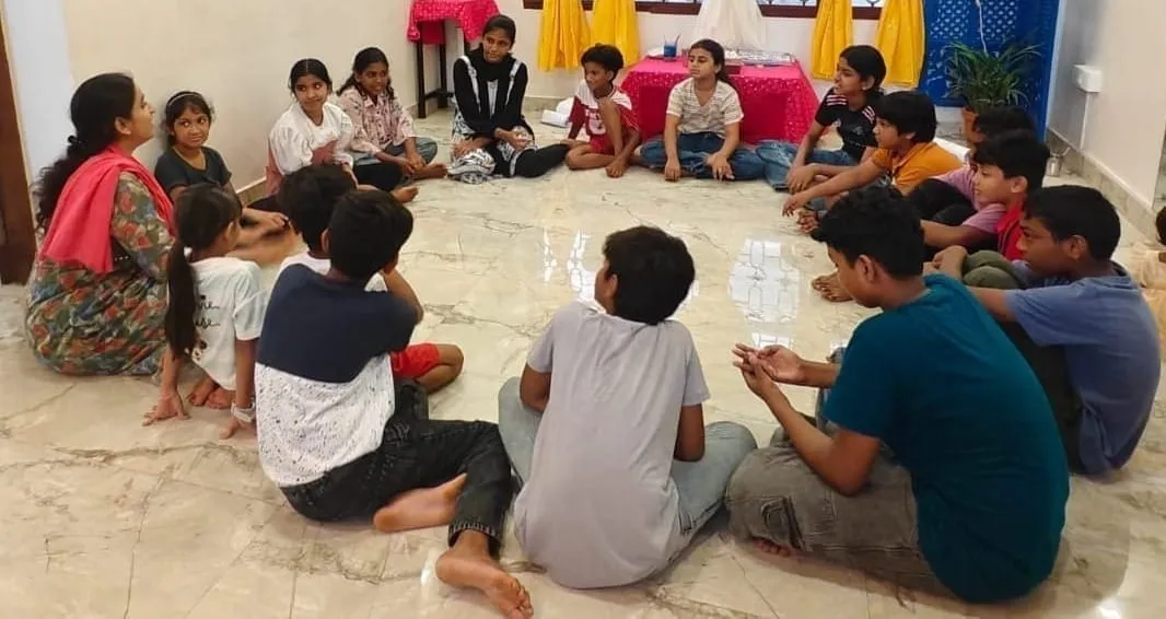 കുട്ടികൾ ഒരു മാർബിൾ തറയിൽ അവർക്കിടയിൽ ചെറിയ പേപ്പർ കപ്പുകളുമായി വട്ടത്തിൽ ഇരിക്കുന്നു, വട്ടത്തിന്റെ അരികിലായി സാരി ധരിച്ച ഒരു മുതിർന്ന സ്ത്രീയുമുണ്ട്.