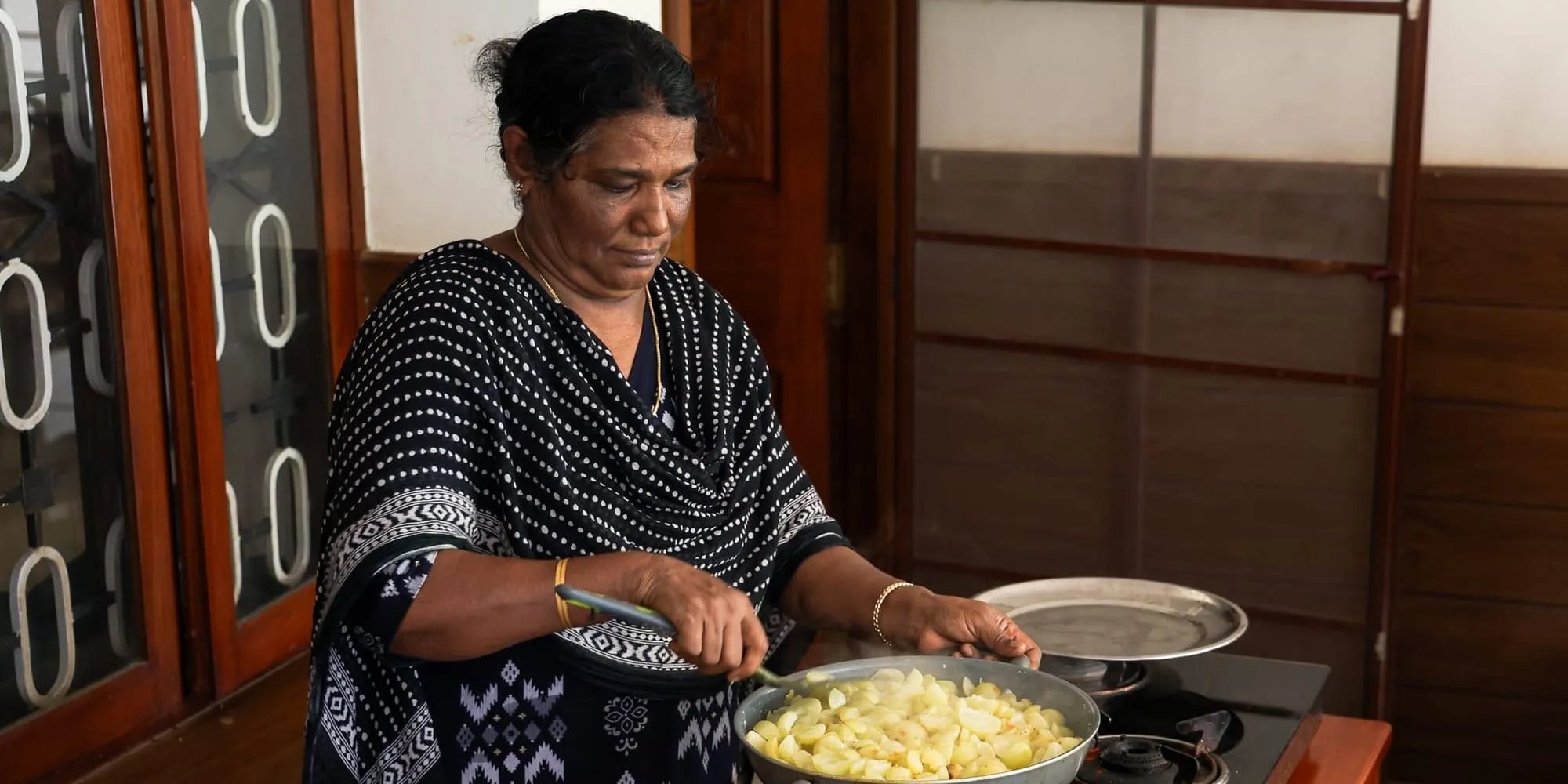 Jasentha Joy stirs a large pan of chopped vegetables on a stove, in a black-and-white patterned top, wood-panelled doors behind her.