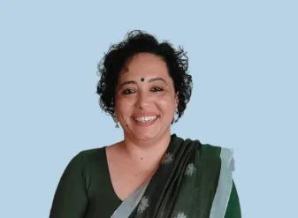 सुमी थॉमस हल्के नीले रंग की पृष्ठभूमि के सामने माथे पर बिंदी और ग्रे बॉर्डर वाली गहरी हरी साड़ी में कैमरे की तरफ देखकर मुस्कुरा रही हैं।