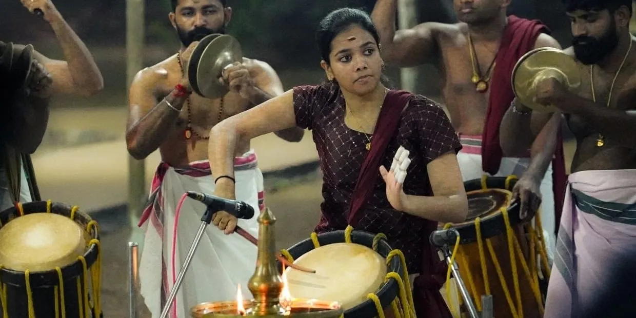 Rahitha Krishnadas plays the Chenda slung at her waist, in a maroon kurta, flanked by men in white mundu playing Chenda and cymbals, a lit brass lamp in front.