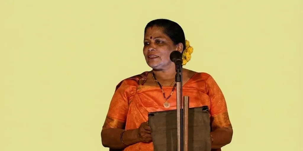 മായ പി എസ് ഓറഞ്ച് സാരിയും മുടിയിൽ പൂക്കളും ചൂടി, വിളറിയ മഞ്ഞ-പച്ച പശ്ചാത്തലത്തിൽ മൈക്രോഫോണുള്ള പ്രസംഗപീഠത്തിൽ സംസാരിച്ചുകൊണ്ട് നിൽക്കുന്നു.