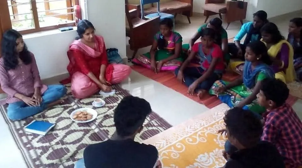 വെളിച്ചമുള്ള ഒരു മുറിയുടെ നിലത്ത് ഡിസൈനുള്ള പായയിൽ കുട്ടികളും യുവതികളും വട്ടത്തിൽ ഇരിക്കുന്നു, അവർക്കിടയിൽ ഒരു പ്ലേറ്റ് പലഹാരങ്ങളും ഒരു നോട്ട്ബുക്കുമുണ്ട്.