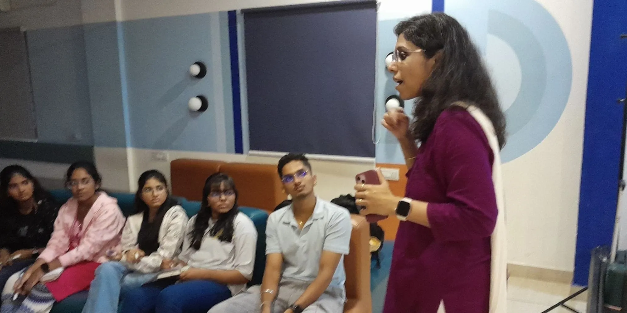स्रीला मेनन मैजेंटा कुर्ते में हाथ से पकड़ने वाले माइक्रोफोन में बोलती हुई खड़ी हैं, सोफे पर युवाओं की एक पंक्ति बैठी सुन रही है, पीछे एक प्रोजेक्टर स्क्रीन है।
