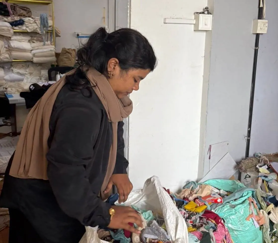 Kadeeja Fidha bends over an open sack of mixed cloth and clothing in a store room, reaching in with one hand, bundles of fabric stacked on shelves behind her.