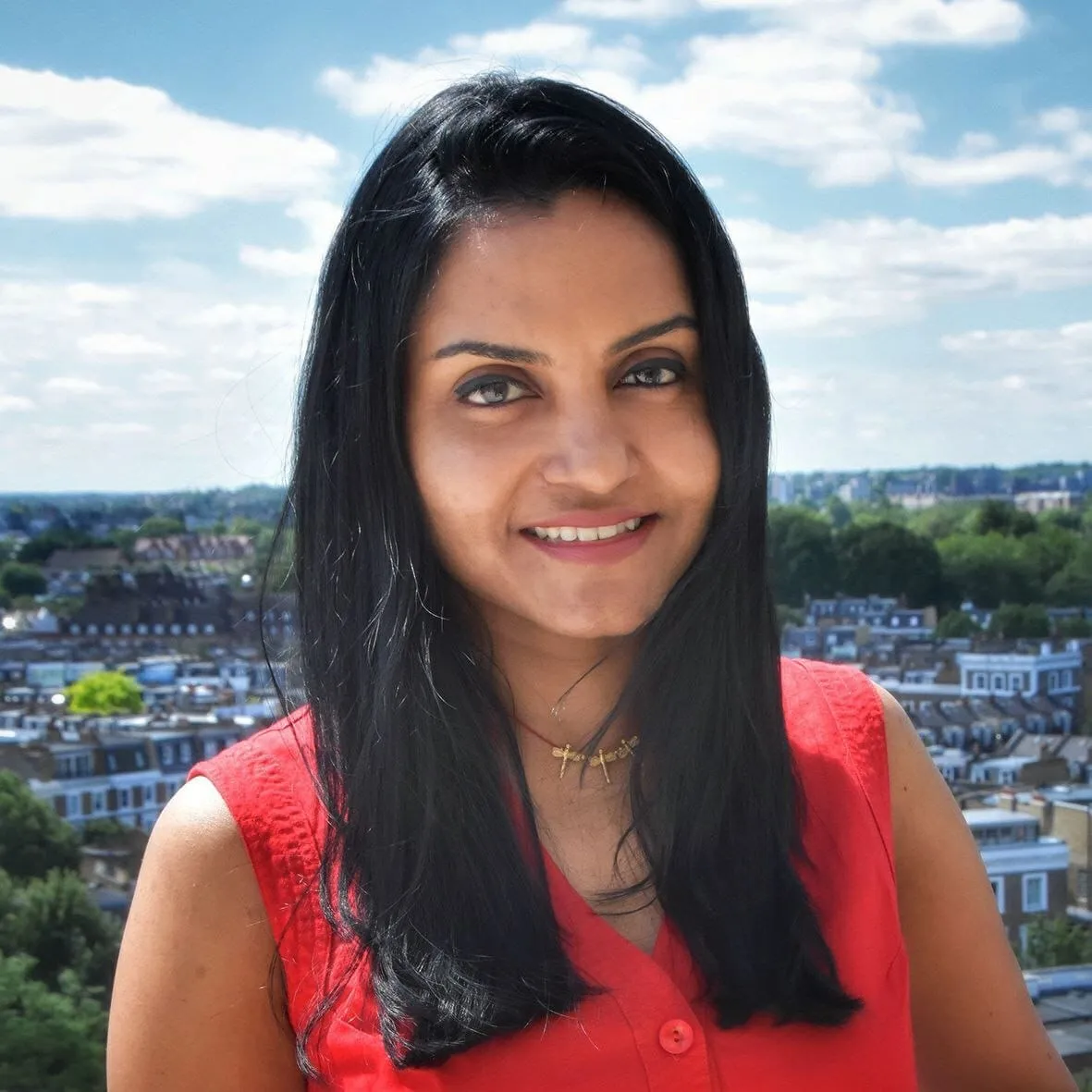 Nidhi Sudhan photographed on a rooftop in a red sleeveless top, city roofs and trees far below and a cloudy sky behind.