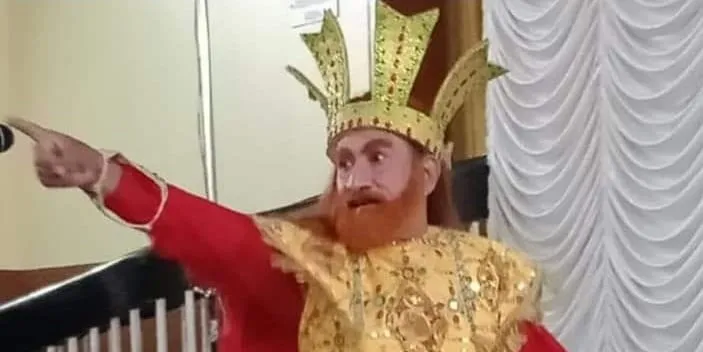 Britto Vincent in a gold crown and a red-and-gold Chavittunadakam costume, one arm flung out mid-scene, performing indoors in front of a white curtain.