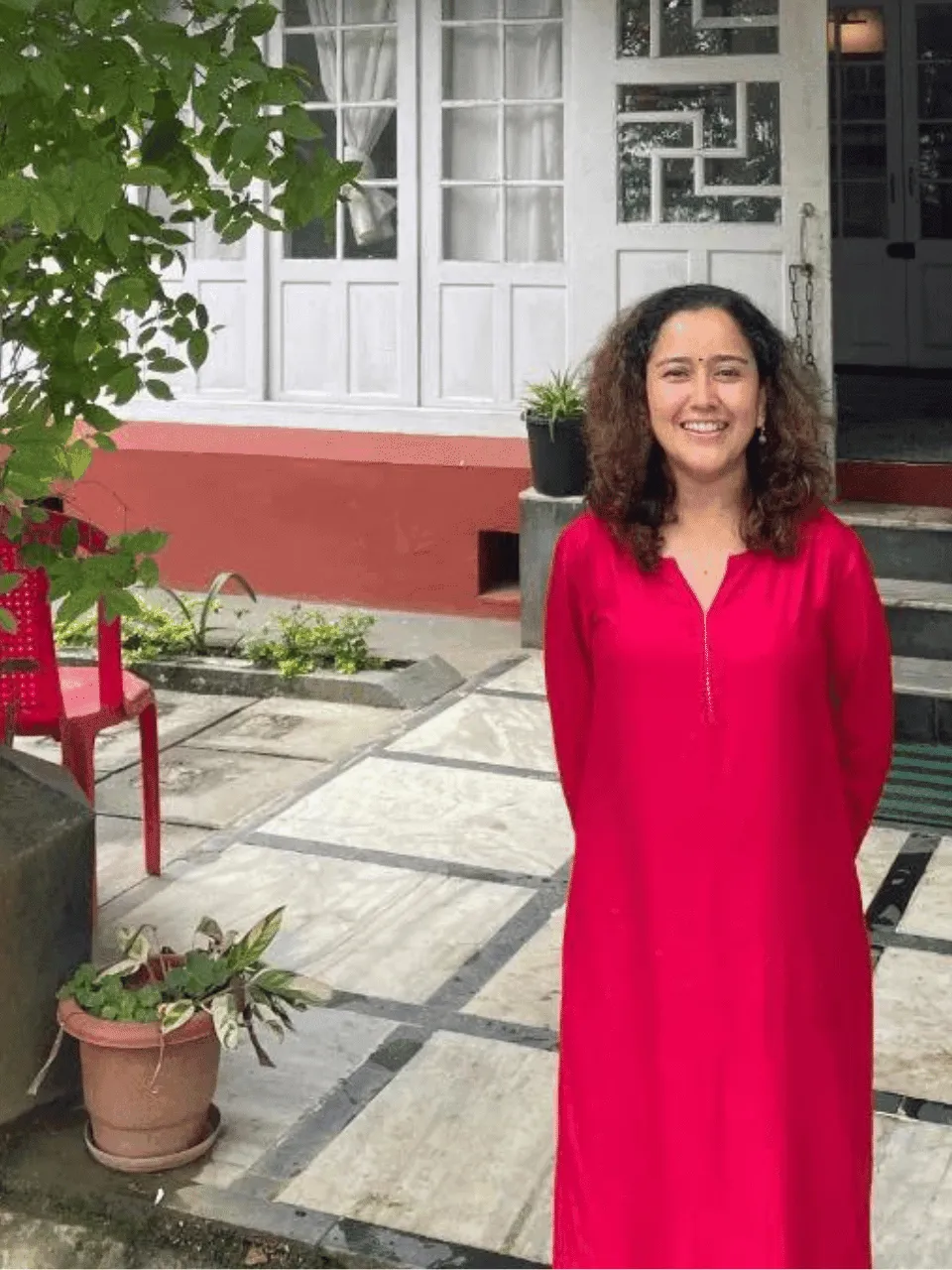Mashqura Fareedi stands smiling on a paved path outside a building with white doors and a red plinth, potted plants around her.