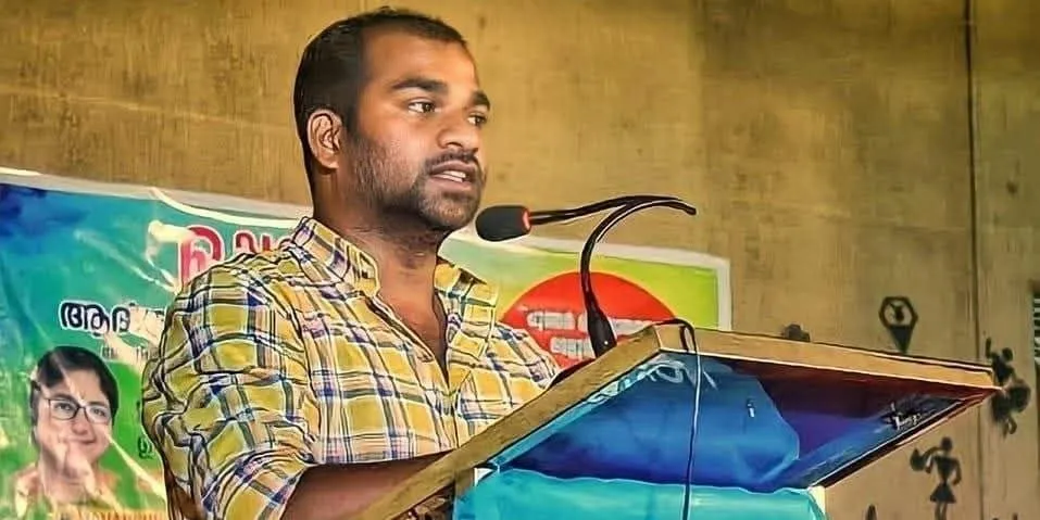 मणिकंदन सी चेक शर्ट पहने एक व्यासपीठ पर माइक में बोल रहे हैं, उनके पीछे मलयालम में एक मुद्रित बैनर है।