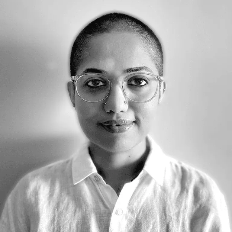 Black-and-white portrait of Shradha in round glasses and a white collared shirt, close-cropped hair, against a plain grey background.